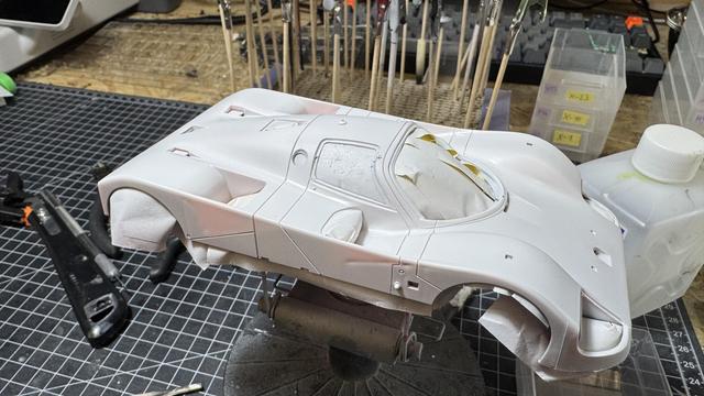 A white-primed model car body mounted on a painting stand with paper towels protecting wheel arches. The workbench beneath shows tools, bottles and a cutting mat.