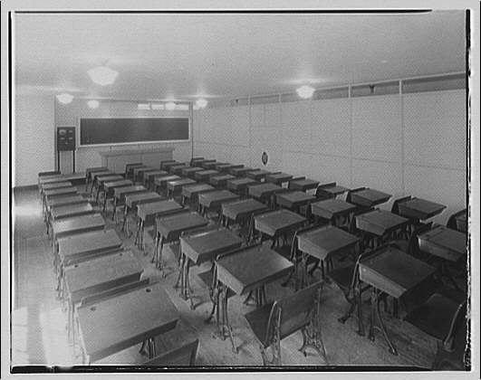 The image depicts a vintage classroom setting, likely from the early to mid-20th century. The room is spacious with multiple rows of wooden desks on high legs arranged in an orderly fashion facing towards what appears to be a chalkboard or projector screen at the far end. Each desk has individual chairs attached underneath. The walls are plain and feature some small round fixtures, possibly for electrical outlets or light switches, as well as a mounted object that could be a clock or sign on one wall.

The classroom is illuminated by overhead lighting with two visible lamps hanging from the ceiling above each row of desks. There's no evidence of any students present in the room at this moment; it appears empty and unused. The image has a black-and-white tone, suggesting an older photograph possibly taken before color photography became widespread or commonly used for such institutional settings.

Overall, the environment reflects a historical educational setting with minimal distractions, focused on teacher-pupil interaction centered around instructional tools like chalkboards or projectors.