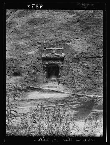 The image shows a black and white photograph of an ancient niche carved into the side of a rock face. The carving appears to be made from stone, with visible layers and texture that suggest it was weathered over time. Above the main opening of the niche is another smaller space containing what looks like shelves or tiers. In front of this scene, there are some plants growing at the bottom edge of the photo, indicating a natural setting for this archaeological site.
The photograph has markings on its borders and corners that resemble an inventory number "V-28" in Arabic script ("م-" in Latin transcription) along with several human figures drawn or written above it. There's also text below depicting what seems to be flora from the area, possibly suggesting a location related to archaeology.
In summary, this image captures a historical site of cultural and archaeological significance, likely situated near Petra (Wadi Musa). The photograph has been attributed to American Colony in Jerusalem Photo Department with one negative measuring 4x5 inches made on nitrate material.