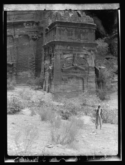 The image is a black and white photograph depicting an ancient rock-cut structure with elaborate carvings. The architecture appears to be of historical significance, possibly from the Greco-Roman era or earlier, given its classical design elements such as columns, friezes, and decorative cornices.

In the foreground, there's a person dressed in traditional Middle Eastern attire, standing on what looks like arid desert ground with sparse vegetation. The individual seems to be observing the structure, possibly indicating interest in it. Behind this figure is an even larger rock-cut building adorned with intricate carvings that depict human figures and decorative motifs.

The photograph has visible borders around its edges, which contain text or annotations providing context about the location and historical significance of the site depicted. The image appears to be from a collection related to American Colony in Jerusalem's Photo Department archives, suggesting it might have been taken during an archaeological expedition or exploration trip circa 1920-1933.

Overall, this photograph provides insight into ancient architecture and offers glimpses into historical research practices of the time.