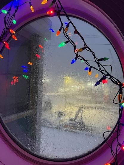 A wintery pre-dawn view through a round window draped with a string of multi-coloured lights. The window is coated with slushy snow, giving the view of the yellowish street lights and machinery a hazy texture.  The window frame glows purple from the reflection of the inside lights, and individual bulbs are reflected multiple times in the window. [St. John’s (NL, Canada) December 2025]