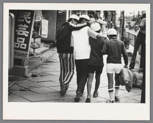 The image depicts a candid street scene, likely taken in Japan given the visible Japanese characters on a signpost. It features four individuals walking together; three of them are embracing each other affectionately from behind while one walks alongside and slightly ahead of them carrying bags over his shoulder.

From left to right: The first person is wearing striped pants with vertical black lines, dark shoes, and appears to have an arm around the shoulders of a second individual. This latter figure has on white footwear, possibly sneakers, and seems to be holding something in their hands that's not clearly visible due to perspective.

The third man shares similar attire as his companion: he wears shorts paired with socks extending up to mid-calf height; however, unlike them, he is wearing a hat backward. His shoes match the others', suggesting they are part of a group or event. The fourth individual walks beside him in white footwear and carries various items such as what seems like a jacket.

The street scene includes pedestrians walking on paving stones with commercial establishments to one side featuring signs that likely indicate local businesses, possibly food-related given their nature.