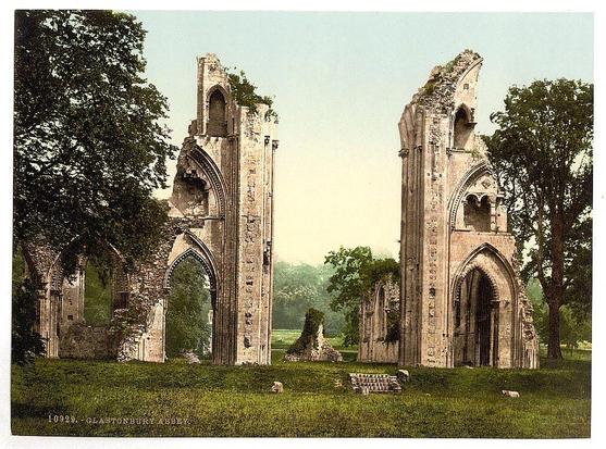 The image shows a pair of ruins from an old abbey. They are likely part of Glastonbury Abbey, as indicated by the text in the lower left corner and confirmed by external sources. These structures have Gothic architectural features such as pointed arches and ribbed vaults within their remaining walls.
Two large pillars stand prominently on either side with intricate stonework including capitals that suggest a Romanesque influence alongside elements of later medieval architecture like the pointed archways, which are indicative of Gothic style. Between these two columns is an open area where green grass covers the ground and trees encircle them, hinting at their historical significance preserved amidst nature.
There's no visible sky in this photo; instead, a soft gradient from pale to lighter hues suggests either dawn or dusk light conditions. Sheep graze peacefully on the lawn within sight of these ruins adding life to an otherwise somber scene of decay and abandonment.
In the foreground lies what appears to be remnants of stone blocks organized neatly into piles which could have once been part of building materials, possibly for repair work that has not yet commenced or perhaps they were simply left behind by earlier renovation efforts. The presence of these stones gives a sense of ongoing human interaction with this historical site.