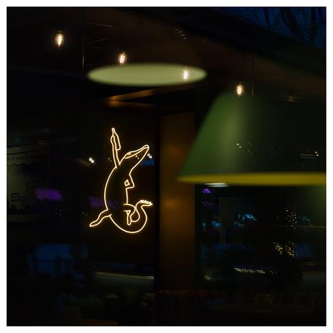 En fyrkantig bild genom en fönster till en folktom restaurang. Till vänster i ljusgul neon hänger en ödla med uppsträckt arm med en öl i handen. Två taklampor i grönt hänger ur fokus i förgrunden. I övrigt diverse små färgglada lampor.