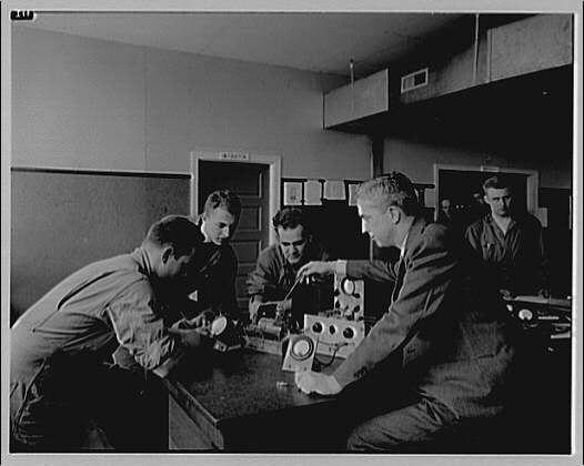 A black and white photograph showing a group of men gathered around radio equipment on tables, with one man pointing at or adjusting the controls. The setting appears to be an office or laboratory environment, possibly related to engineering or electrical work during the early 20th century.
This image captures students and their teacher examining various pieces of vintage radio-related machinery in what seems like a professional educational setting from around 1920-1950.