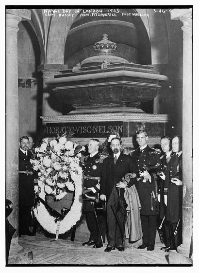 The image is a black-and-white photograph from 1923, capturing a formal event labeled as "Naval Day in London." The setting appears to be inside a grand, ornate building with arched ceilings and stone columns, suggesting a significant historical or ceremonial location. In the center, there is a large, ornate monument with a crown-like structure on top, which is likely a memorial or monument. The text on the monument reads "HORATIO VISC NELSON," indicating it is dedicated to Horatio Nelson, a famous naval hero.

In front of the monument, a group of individuals is gathered. They are dressed in formal military attire, suggesting their involvement in the naval or military sphere. Some are wearing uniforms with medals and decorations, and others are in civilian suits. One individual is holding a large floral wreath, which is placed in front of the monument, symbolizing respect or commemoration. The individuals are standing in a line, with some holding swords, adding to the ceremonial atmosphere. The photograph has a caption at the top that reads "NAVAL DAY IN LONDON 1923," along with names and titles, such as "CAPT. HUSSEY," "ADM. FITZMAURICE," and "POSTWHEELER," which likely refer to the individuals present in the photograph. The image is marked with a number "3146-9," possibly indicating it is part of a collection or archive.