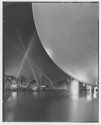 The image depicts a nighttime scene featuring the Perisphere, an iconic structure from the 1939 World's Fair held in New York City. The photograph captures the spherical shape of the building with its smooth and expansive surface reflecting light from multiple beams that converge at its apex. These lights create striking patterns on both the perisphere and the surrounding water body.

The sky is dark, indicating it is nighttime, which adds a dramatic contrast to the bright lighting effects emanating from within or around the structure. The overall composition of the image highlights the grandeur and architectural beauty of this landmark as part of an exhibition meant to showcase modern engineering feats during that time period.