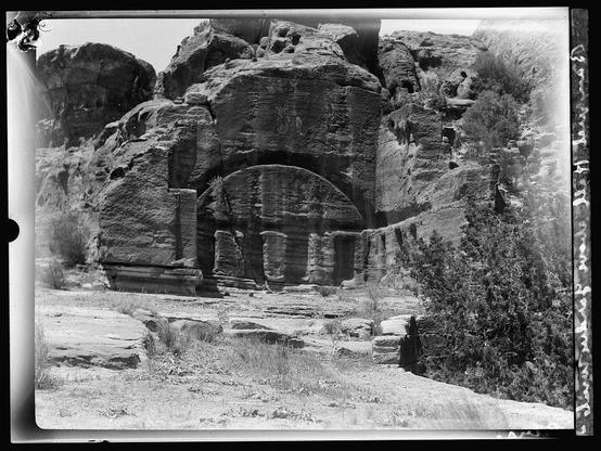 The image depicts an ancient rock-cut architecture, likely a historical site with significant cultural and archaeological value. The structure appears to be carved into the side of a cliff or rocky outcrop, featuring multiple levels and archways that suggest it was once part of larger buildings or complexes.
In the foreground, there's arid landscape with sparse vegetation, indicative of a dry climate region such as Wadi Farasah in Petra. The absence of people provides an unobstructed view of the intricate stone workmanship.
The black-and-white nature and grainy texture suggest it is likely from historical photographic records, possibly around 1920 to 1933 based on the caption which references Roman hall periods and specific location details like El-Farasah & obelisk ridge area. The image might have been taken by American Colony (Jerusalem). Photo Department as per a negative of nitrate material indicating its age-old origin.
Further context can be found in Petra, an ancient city known for its unique rock-cut architecture and historical significance dating back to the Nabatean civilization around 200 B.C.