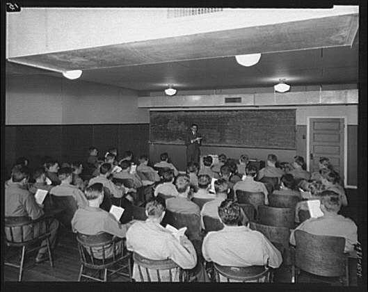 The image depicts a classroom scene where numerous individuals are seated and facing the front, presumably attending a lecture. The room is equipped with several rows of chairs filled by students wearing what appears to be military-style uniforms or casual attire. Each student holds an open booklet in their hands, indicating that they might have materials for note-taking.

In the center stands a person at the forefront of the classroom, positioned behind a lectern, addressing the audience. This individual is likely the instructor conducting the lecture. The front wall features a large chalkboard filled with writing and diagrams, suggesting an educational or instructional context.

The setting appears to be indoors with fluorescent lighting overhead from ceiling fixtures. There are no windows visible in the picture, reinforcing that it's taken inside a building designed for teaching purposes. Overall, this image captures a moment of learning within an organized academic environment likely associated with military training during the early 20th century based on attire and equipment used by students.

Please note that additional information about this specific photograph can be found in Capitol Radio Engineering Institute archives or records from approximately ca.1920-ca.