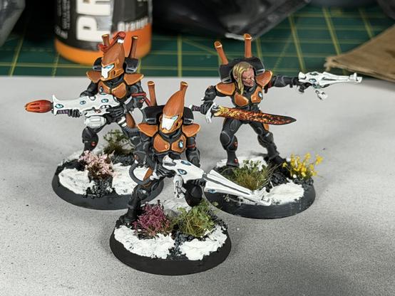 Three Games Workshop Eldar Guardians, painted grey and orange in the scheme of Yme-Loc, with white weapons on a snowy base. The front guardian is carrying a shuriken catapult. The rear left guardian is carrying a fusion gun, which has a fiery red tip. The rear right guardian is carrying a sword and shuriken pistol, the sword is flaming. They are also not wearing a helmet.