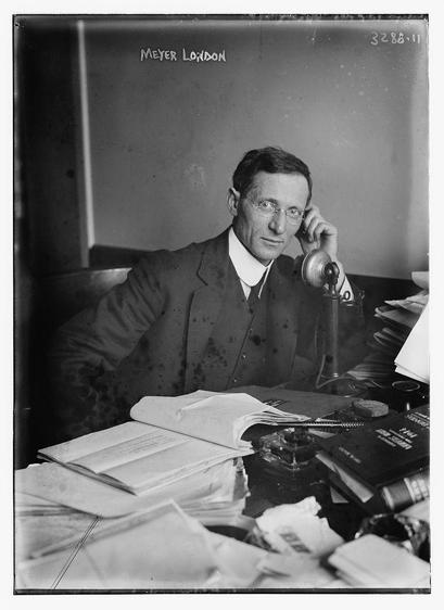 The image is a black-and-white photograph of a man seated at a cluttered desk. He is wearing a suit and tie, and his attire suggests a formal or professional setting. The man appears to be in his middle age and is wearing glasses. He is holding a telephone receiver to his ear with his left hand, and his expression is neutral, with a slight tilt of his head. The desk in front of him is filled with papers, books, and what appears to be a desk lamp, indicating a busy work environment. The background is plain and dark, which draws attention to the man and the desk. There is text at the top of the image that reads "MEYER LONDON," and a number "3288.11" is visible in the upper right corner. The overall style of the photograph and the attire of the man suggest it is from the early 20th century.
