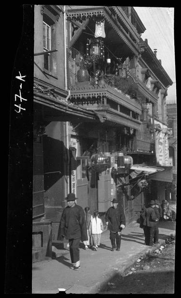 The image depicts a street scene, likely from Chinatown in San Francisco during the late 1800s or early 1900s. The architecture is characterized by ornate painted balconies with intricate details and designs that suggest an Eastern influence, possibly Chinese. These structures are multi-storied buildings adorned with hanging plants, lanterns, and various decorative elements.

The street below shows a bustling scene with several individuals walking along the pavement. Some of these people appear to be wearing traditional East Asian attire, including hats known as "ding ding," which were commonly worn by men in that era for sun protection or fashion purposes.

The overall atmosphere conveys a busy urban environment with historical significance due to its architectural style and cultural elements. The black-and-white nature of the photograph adds an authentic vintage feel, capturing a glimpse into daily life during this time period.