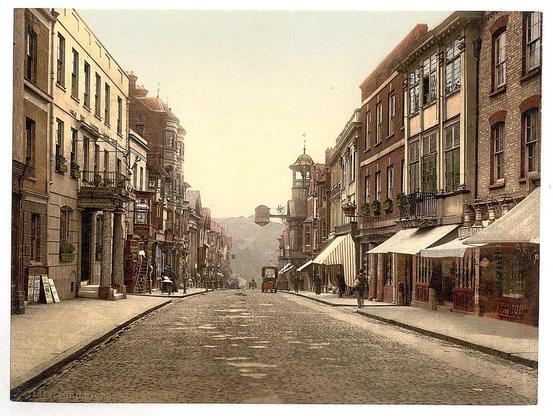 This image depicts a historic urban street scene from what appears to be late 19th century, judging by the architectural style and design of vehicles. The High Street in Guildford is lined with multi-story buildings exhibiting characteristics typical for that era such as ornate facades, large windows, fire escapes on some structures, and visible signage indicating businesses or residences.
The street itself has a cobbled surface, indicative of pre-modern paving methods common before the widespread adoption of modern roads. A horse-drawn tram is in motion down this street, with its single horse pulling an attached carriage-like structure carrying passengers.
On either side of the street are storefronts and residential buildings; some have awnings extending over the sidewalk where pedestrians walk. People can be seen walking along these sidewalks, suggesting a lively urban environment even at that early period. The lighting suggests it could be late afternoon or early evening given the warm hue cast on the architecture.
The overall composition of this image captures daily life and commerce in what seems to be an upper-middle-class district during Guildford's historical heyday before modernization swept through many British towns, signaling a shift towards more industrialized cities.