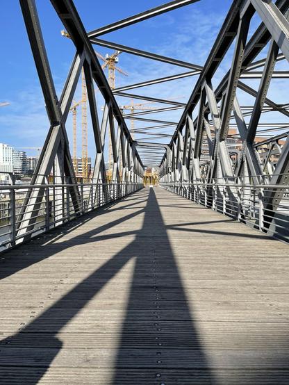 A pedestrian bridge with a geometric metal truss structure, viewed on a sunny day. The bridge deck is wooden with visible rivets and elongated shadows cast by the truss. In the background, construction cranes and modern buildings are visible under a clear blue sky.