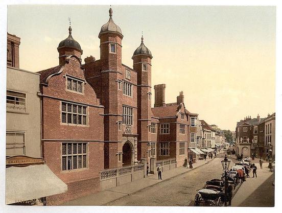 The image depicts a historic street scene in what appears to be the early 19th century. The focal point is an imposing brick building with Gothic architectural features, including two prominent towers capped with domed roofs and conical spires. The structure has several large windows framed by decorative masonry work and gabled dormer windows on its upper story.
The street in front of this building shows a bustling town atmosphere. People are walking along the sidewalk or gathered near storefronts that have awnings. A few individuals appear to be carrying goods, possibly vendors selling items from carts parked alongside the road.
Vintage automobiles and horse-drawn carriages share the narrow roadway, suggesting the transitional period between modernization and traditional modes of transportation. Street lamps line the street at regular intervals, casting a soft glow over the scene.
The sky is hazy with no distinct features such as clouds visible in this sepia-toned photograph, adding to its aged appearance. The overall impression captures the essence of daily life during that era in Guildford, England.
