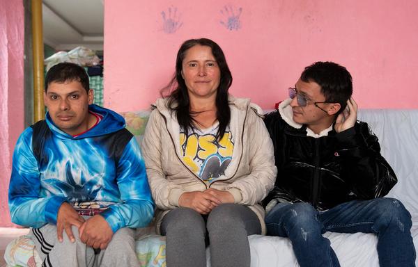 A woman and her two adult sons sit close to one another on a couch covered with blankets. Two purple hand prints decorate the bright pink wall behind them.