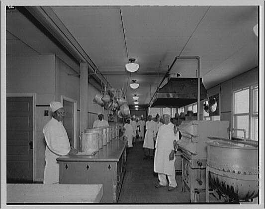 The image depicts a vintage kitchen setting, seemingly from the early to mid-20th century. It is an interior view of what appears to be a professional or institutional cooking area with several individuals in chef uniforms and headgear actively engaged in food preparation tasks.

In this black-and-white photograph, multiple people are seen wearing white coats and hats typical for kitchen staff during that era. They stand at workstations equipped with large stainless-steel countertops which house various utensils like mixing bowls, ladles, and other cooking tools.

Hanging pots can be observed suspended from a rack above the preparation area. The background shows additional equipment such as what appears to be an industrial-size pot or kettles on the right side of the frame. There are also visible overhead fluorescent lights illuminating the space, with some light fixtures being off-camera.

The setting suggests that this could be part of a larger establishment like a hotel, hospital, or perhaps related to food engineering as indicated by the caption "Kitchen at Capitol Radio Engineering Institute." The photograph captures an atmosphere indicative of early industrial culinary practices.