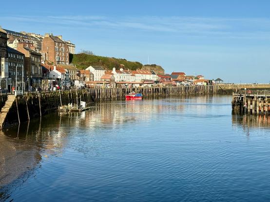 A quiet harbour with calm water reflects a line of historic buildings with brick and stone facades, some topped with red-tiled roofs, stretching along a wooden quay. A few small boats, including a bright red vessel, are moored in the sheltered water, while wooden pilings and sea walls frame the harbour. On the left, a row of shops and houses sits close to the water’s edge, with steps leading down to the quay, and a gentle slope of green hills rises behind the town. Clear blue skies and sunlight highlight the contrast between the calm water and the bustling waterfront.