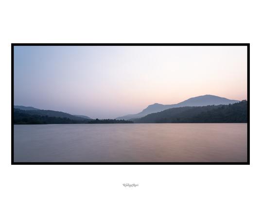 A serene landscape scenery of a lake reflecting the early morning colors - blues, oranges and reds. Rolling hills in the distance.