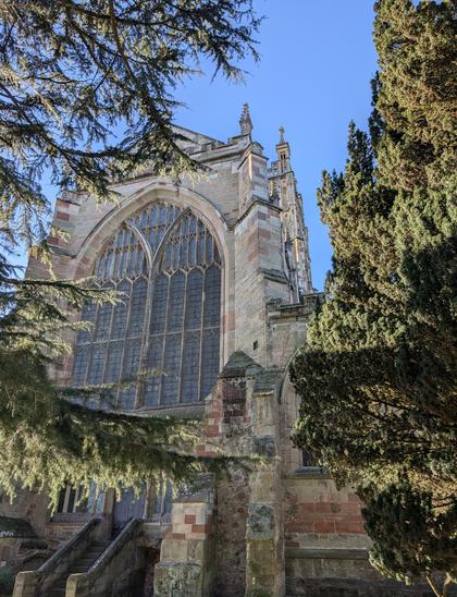 A view of the east end of a large parish church, framed on either side by green foliage. The winter sun is bright and the sky beyond is cloudless blue.