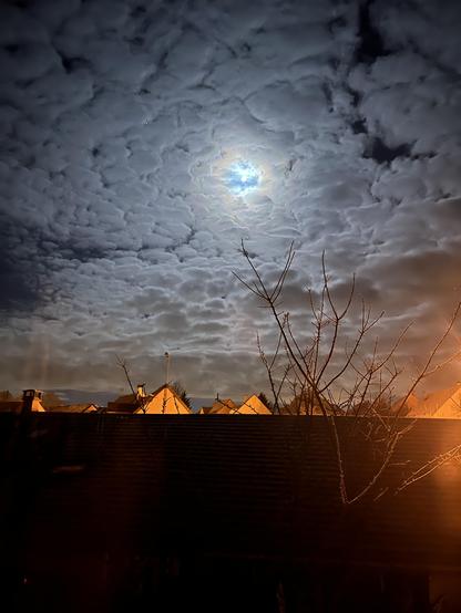 Photographie d’un ciel moutonneux avec la lune toute ronde apparaissant au milieu des nuages. Dans la partie basse de l’image des toits rouges orangés éclairés par les candélabres de la rue.