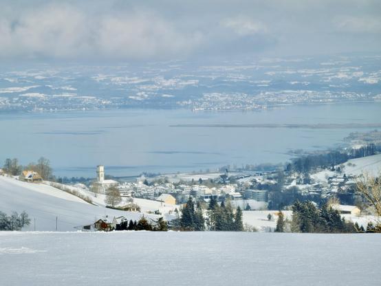 Eine Winterlandschaft mit einem schneebedeckten Hügel mit Blick auf einen ruhigen See, der von fernen Hügeln umgeben ist. Die Szene umfasst malerische Häuser und einen Kirchturm im Schnee, mit einem weichen, bewölkten Himmel darüber.