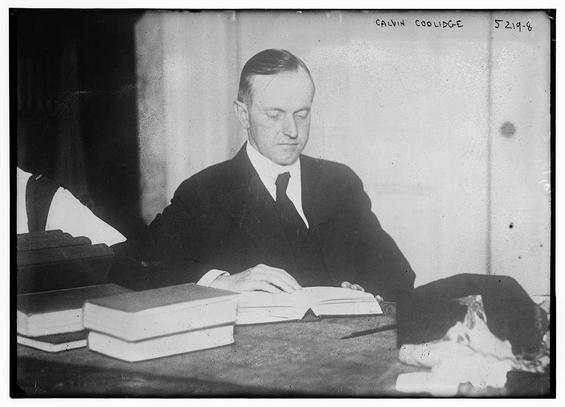 The image is a black-and-white photograph of a man sitting at a desk. He is dressed in formal attire, wearing a dark suit, white shirt, and a dark tie. The man appears to be middle-aged and has short, neatly combed hair. He is seated with his hands resting on an open book, which is placed on the desk in front of him. The desk also holds several other books stacked neatly beside the open book. The background is simple and somewhat dimly lit, suggesting an indoor setting, possibly an office or study. The photograph has a label in the upper right corner that reads "CALVIN COOLIDGE" and a number "5219-8" beneath it. The overall tone of the image conveys a sense of formality and intellectual engagement.