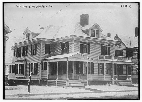 The image is a historical black-and-white photograph of a large two-story house. The house is labeled as "Coolidge Home, Northampton," as indicated by the handwritten text at the top of the image. The architectural style suggests it is from the late 19th or early 20th century, with a symmetrical design and a prominent front porch supported by wooden pillars.

The house features a gabled roof with multiple windows on each floor, some with shutters. The second story has a small dormer window on the left side. The front porch extends across the entire width of the house, supported by evenly spaced wooden columns. The porch has a railing and steps leading up to the entrance.

The house is surrounded by a paved area in front, with a small patch of snow visible on the ground, indicating a winter setting. There are utility poles and wires running along the street in the foreground. The overall scene gives a sense of a residential neighborhood with a well-maintained property.