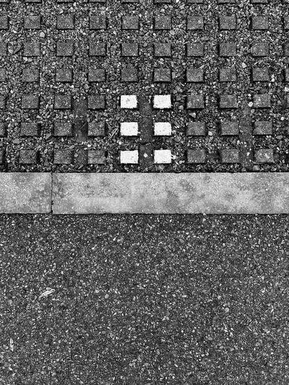 A monotone image with a gravelly surface divided in two horizontally by a cement strip. The top half has a pattern of raised squares, with six squares (in three rows of two) painted white. This is a close-up of curiously subtle parking bay indicators. Brook Drive, Green Park, Reading, Berkshire, UK. Photo by Gearóid Burke.