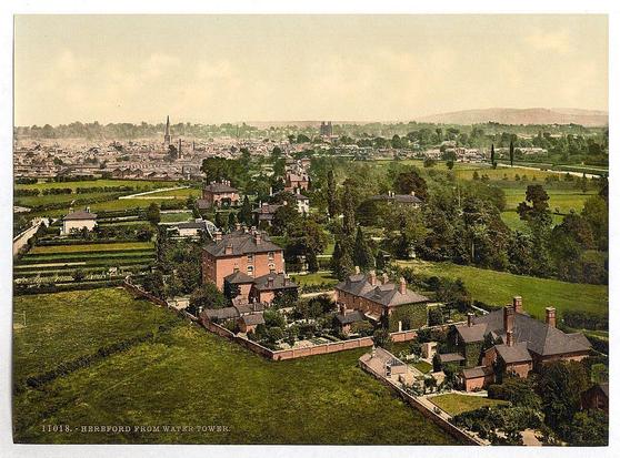 This image is a vintage photograph depicting an aerial view of Hereford, England. The date range associated with the photo suggests it was taken between approximately 1890 and 1900. It's captured from Water Tower in Hereford as indicated by text at the bottom left corner.

The scene shows densely packed urban structures that form a sprawling cityscape extending into the distance. Central to this landscape is an impressive spire, possibly indicating historical or religious significance within the town center. The skyline includes notable buildings with pointed rooftops and chimneys, indicative of residential architecture from the period.

In contrast to the bustling townscape, lush green fields and gardens are visible in the foreground and middle ground. This juxtaposition highlights a mix of urban development and rural expanse typical for British countryside areas during this era. The colors appear somewhat faded but convey an impressionistic quality characteristic of early color printing methods such as photochrom.

The photograph is credited to Loener, suggesting it was produced by or distributed through the Dutch firm known for its photographic work in Europe during that period.