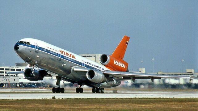 The image shows a wide-body, three-engine jet airliner in the act of landing on a paved runway at Miami airport. The aircraft is a McDonnell Douglas DC-10 operated by VIASA (Venezolana Internacional de Aviación, S.A.), Venezuela’s former flag carrier. The airplane is captured in a left-to-right profile view, slightly nose-up, with its main landing gear already in contact with the runway and producing visible white tire smoke from touchdown friction.

The fuselage is predominantly white on the upper section, with a polished metallic silver lower half. Along the forward fuselage, the airline name “VIASA” appears in large, bold red letters, while “VENEZUELA” is written in blue along the midsection. A row of passenger windows runs the length of the cabin. The vertical tail is painted a vivid orange, prominently marked with the airline’s name “VIASA” in white block letters, and a small Venezuelan flag near the top.

The DC-10’s distinctive engine configuration is clearly visible: two large turbofan engines mounted under the wings and a third engine integrated into the base of the vertical tail, with its intake visible at the tail root. The wings extend outward with flaps fully deployed, indicating landing configuration. The nose gear is extended but has not yet fully settled onto the runway.

In the background, airport terminal buildings and ground infrastructure are visible, rendered slightly out of focus, suggesting a busy international airport environment.