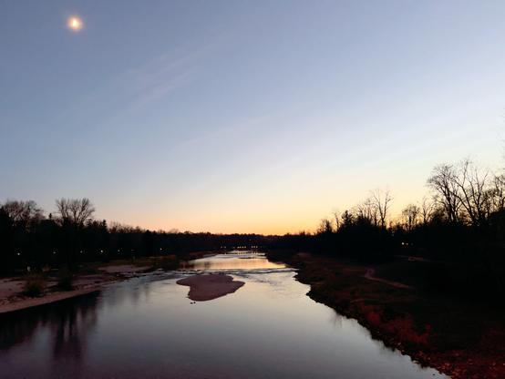 Eine ruhige Flussszene in der Dämmerung, mit einem teilweise sichtbaren Mond am Himmel. Der Horizont wird von einem Sonnenuntergangsglühen beleuchtet, und Bäume säumen die Ufer. Ruhiges Wasser spiegelt die Farben des Himmels wider, mit einer kleinen Sandinsel im Fluss.