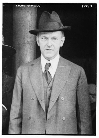The image is a black-and-white photograph of a man standing in front of a dark background. The man is wearing formal attire, which includes a double-breasted suit jacket, a white dress shirt, and a dark tie. He is also wearing a fedora-style hat. The image appears to be a portrait, and there is text at the top left corner of the photograph that reads "CALVIN COOLIDGE." The style of the photograph and the man's attire suggest it is from the early to mid-20th century. The lighting is even, and the subject is centered in the frame. The man's expression is neutral, and his posture is straight, giving the impression of formality and respectability.