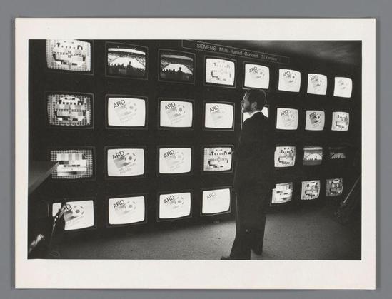 The image depicts a man standing in front of an array of television screens. He is dressed formally, wearing a suit and tie, which suggests that he could be a professional or possibly part of the media industry. The TVs display various channels with the logo "ARD," indicating they are likely broadcasting German content from Deutsche Gesellschaft für Fernsehen und Rundfunk (ARD), Germany's public-service broadcaster.

The man appears to be observing one screen closely, which shows what seems like footage on a sports field, possibly related to an ongoing match. The array of screens is part of a large installation, as it extends beyond the frame and suggests that this could be in a control room or newsroom setting where multiple streams are monitored simultaneously.

The black-and-white aesthetic gives the photo a vintage feel, indicating it was taken at least several decades ago when color photography had not yet become ubiquitous. The caption "SIEMENS Multi-Kanal - Kanaal-Concept" hints that Siemens developed this system for monitoring various channels in parallel. This technology reflects advancements of its time and signifies how media consumption has evolved from single-channel viewing to multichannel broadcasting.

The photo is credited with a reference URL, which likely leads to more information about the project or context within which it was captured by Ed van der E [...]