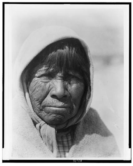The image is a black and white photograph, showing an elderly Indigenous woman wearing what appears to be a fur hood or head covering. Her face shows signs of aging with deep wrinkles and fine lines around her eyes, nose, and mouth. She has dark hair that seems to peek out from under the edge of the hat. The background behind her is blurry but suggests she may be outdoors in an open space.
The woman appears contemplative, possibly lost in thought or simply looking into the distance. Her expression conveys a sense of weariness yet also resilience and perhaps introspection. She's wearing what seems to be layered clothing that indicates it might be cold weather; her attire could suggest cultural significance as well. The image is presented on white paper with some text at the bottom, likely indicating metadata or additional information about this photograph.
This particular photo carries a historical context since it was taken by Edward S. Curtis (1868-1952), an American photographer known for his work documenting Native Americans in North America during the early 20th century. The title "Sù̂-Donii ("Osier-Willow Blossom")--Pyramid Lake Paviotso c1924" suggests that this photograph is part of Curtis' extensive body of work on indigenous peoples, possibly captured around Pyramid Lake in Nevada during the 1920s. The image serves as a visual record and cultural heritage from th [...]
