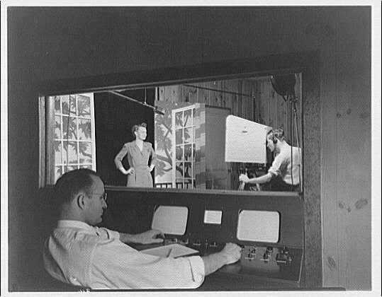 The image depicts a vintage television studio scene from approximately the year 1945. A man is operating control panels with numerous buttons and dials, indicating he's likely managing various aspects of live broadcast technology during that time period. In front of him in a reflective mirror is a woman standing confidently with her hands on her hips; she appears to be an actress or presenter who has stepped out from the set. Another man stands behind equipment connected to microphones and cables, possibly involved in sound recording or production tasks. The image captures the early days of television broadcasting technology as it was developed at Capitol Radio Engineering Institute VI during that era.