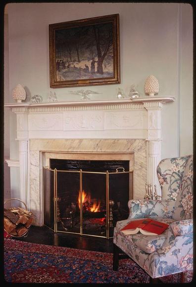 This image features a warm and inviting living room with an emphasis on classic design elements. At the heart of the space is a white marble fireplace mantel, adorned with various decorative items such as glass vases, pine cones, and candlesticks. Above the fireplace hangs a framed painting depicting a winter scene.

In front of the mantel stands a brass fireplace screen that partially obscures the view of the fire within it. Adjacent to the mantlepiece is an upholstered armchair decorated with floral patterns in shades of blue and pink on a white background, suggesting a vintage or antique style. A red book rests casually across one armrest.

A vibrant Persian-style rug covers part of the hardwood floor underfoot, adding warmth and color to the room's overall aesthetic. The surrounding walls are painted a neutral light beige tone that enhances the cozy atmosphere without drawing attention away from the central fireplace feature.

This image is likely drawn from Toni Frissell’s photograph collection for Vogue magazine, which showcases American fashion photography during the 1960s-1970s era with notable portraits of prominent individuals like Gracie Mansion. The photo was published in May 1969 and can be found within Details Magazine's coverage.

The online repository at Library of Congress provides further information about Frissell’s work, which spans various genres incl [...]