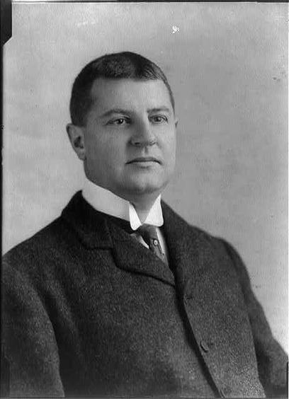 The image is a black-and-white portrait of a man dressed in formal attire, likely from the early 20th century. The man has short, neatly combed hair and is looking directly at the camera with a neutral expression. He is wearing a dark suit jacket with a high collar, a white dress shirt, and a dark tie. The background is plain and light-colored, which helps to emphasize the subject. The overall style of the photograph and the man's attire suggest it could be from the late Victorian or Edwardian era. The lighting is even, and there are no visible distractions in the background.