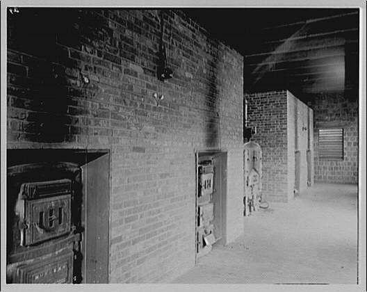 A black and white photograph showing the interior of a basement room with brick walls, including several doors or compartments. The left side features an old-fashioned furnace, while on the right wall there are various utility fixtures such as electrical boxes, possibly for wiring purposes. A figure is faintly visible in the distance standing beside these utilities.