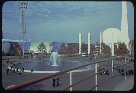 The image depicts an outdoor scene at a World's Fair, specifically the Plaza of Light. In the center is a fountain with water jets shooting upwards, creating a circular pattern on the surface below it. Surrounding this focal point are several tall, white pillars and a large, cylindrical structure that appears to be part of exhibition buildings or landmarks within the fairground.

The plaza seems spacious with people leisurely walking around and others seated in groups near the fountain's edge. The sky is clear, suggesting pleasant weather conditions during the time the photo was taken. In the background, there are more architectural elements including a large white dome-like structure on one side which might be part of an exhibition hall or another significant building at this fair.

The image appears to have been captured by Gottscho-Schleisner Inc., as indicated in the credit information provided for this particular slide. The reference "World's Fair Plaza of Light, toward Theme Center 1939 or 1940" suggests that this photograph was taken at an event likely held around those years and features a specific area within the fair known as the 'Plaza of Light.'