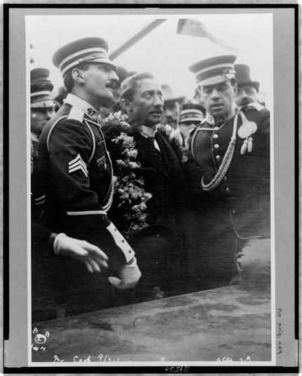 The image is a black-and-white photograph featuring a group of individuals, likely from the early 20th century, based on their attire and the style of the photograph. The individuals are dressed in formal military and civilian uniforms, suggesting a formal or ceremonial event.
In the foreground, a person wearing a military uniform with a cap and epaulets is prominently visible. The uniform includes a number "47" on the sleeve, indicating a specific rank or unit. This individual appears to be interacting with others, possibly greeting or conversing with them.
To the right of the military figure, there is a man in a dark suit with a bow tie, who seems to be part of the group. He is looking to the side, possibly engaging with someone out of frame. Behind him, another individual in a military uniform with medals and decorations is visible, indicating a high-ranking or distinguished person.
The background is crowded with other individuals, some also in uniform, suggesting a gathering or public event. The photograph has a caption or notation in the bottom left corner, which reads "Dr. Carl 9/13/13," and in the bottom right corner, there is a notation "no. orig. neg." which likely refers to the original negative of the photograph.
The overall atmosphere of the image suggests a formal occasion, possibly a military or diplomatic event, given the attire and the presence of individuals in uniform.