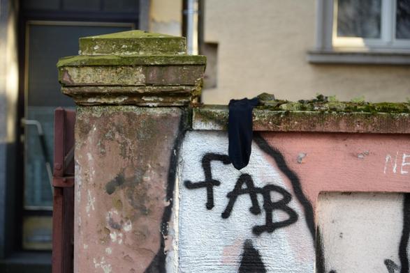 Eine schwarze Socke hängt über einer Mauer mit Pfosten. Darunter auf der Mauer ist als Teil eines Greaffitis der Schriftzug FAB zu sehen.
