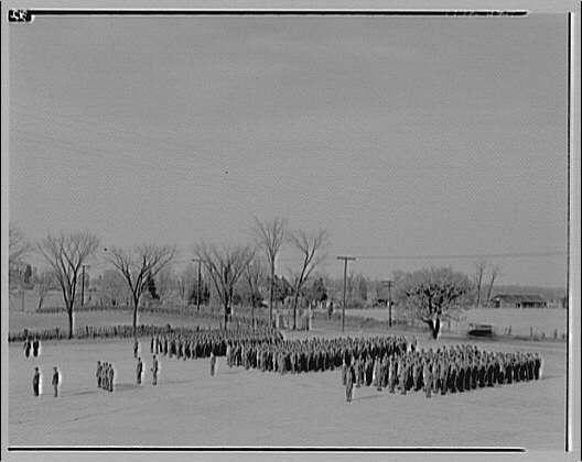 The image depicts a large group of uniformed men standing outdoors, arranged in what appears to be a formation for a review or inspection. The scene is black and white, suggesting it might be from the early 1900s based on the attire and style of photography.
In this historical photo captured around ca. 1920-ca. 1950 at Capitol Radio Engineering Institute by Horydczak, a notable photographer who lived approximately between 1890-1971, we see rows upon rows of men dressed in military-style uniforms standing on what looks like an expansive field with barren trees and distant buildings visible under a clear sky. The ground seems to be snow-covered or at least very dry, which may indicate winter time.
Several individuals stand apart from the main formation: some near the front left corner appear slightly out of alignment as if walking towards or away from their positions in line; others on either side seem more relaxed and might not have been formally included. The overall atmosphere is one disciplined organization within an outdoor setting, typical for military or government-related activities during that era.
The image has a caption indicating its source - Capitol Radio Engineering Institute with the photographer's name Horydczak mentioned as well along with their lifespan range (1890-1971). The black and white nature of this photograph serves to focus on details like uniform [...]
