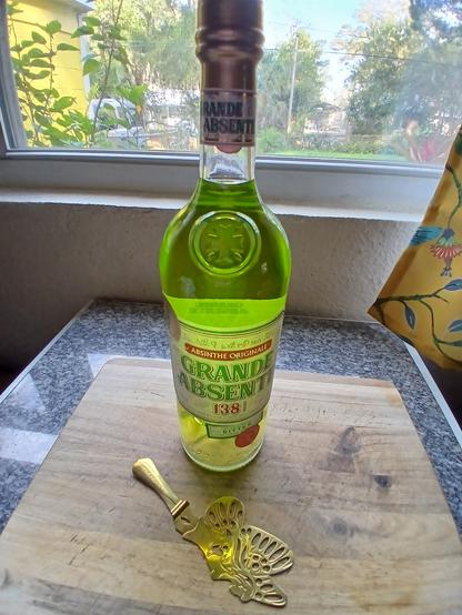 A bottle of absenthe and green fairy spoon on a cutting board.