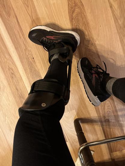 POV,  looking down at legs in black leggings. Left leg wears a wrap around prosthetic that is newly refurbed and reshaped that fastens under the knee, carries movement down the back of the calf on long posts and into supports that frame the ankle and continue into running shoes. It’s carbon fiber exterior with mechanisms for fastening and soft foam inside