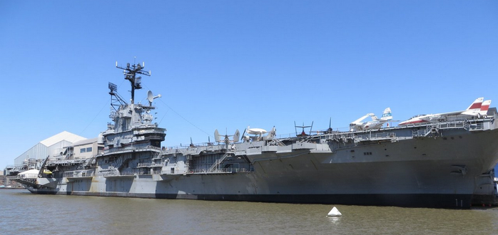 Ein großer grauer Flugzeugträger liegt in einem Hafen unter einem klaren blauen Himmel. Die Schiffs-Flugdeck zeigt mehrere Flugzeuge, darunter ein weißes Flugzeug, ein blau-weißes Flugzeug und ein rot-weißes Flugzeug. Die Schiffs-Überhänge verfügen über Antennen und Radargeräten. An der linken Seite ist ein Gebäude mit einem weißen Dach sichtbar, nahe dem Schiff. Das Wasser ist ruhig und braun, mit einem kleinen weißen Schiffsbau in der Nähe.