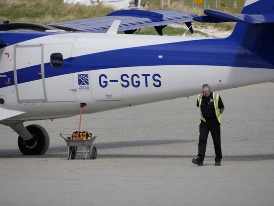 Ein blau-weisses kleines Flugzeug (Twin Otter) auf einem Strand. Ein Mann mit Warnweste steht daneben. In einer Schubkarre neben dem Flugzeug ist ein Generator der das Flugzeug mit Strom versorgt.