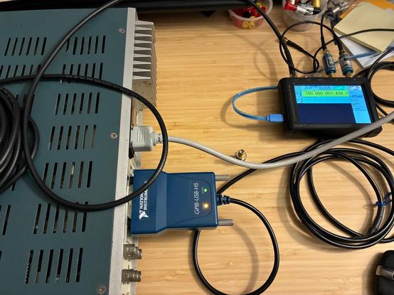 Desktop photo from above showing a NI GPIB-USB-HS module connected to the rear of the Fluke 1985A counter. Coaxial cable from the TinyGTC clock output is routed to the counter's A input jack.