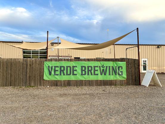 Verde Brewing sign on a green banner on the wooden fence under the partly sunny sky.