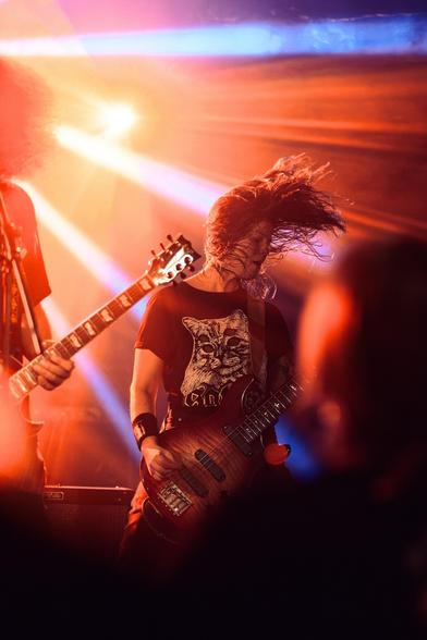 A musician is performing on stage, energetically playing a bass guitar while headbanging. Bright stage lights create a colorful, dynamic atmosphere. Another guitarist is partially visible in the background. The scene conveys a lively concert vibe.